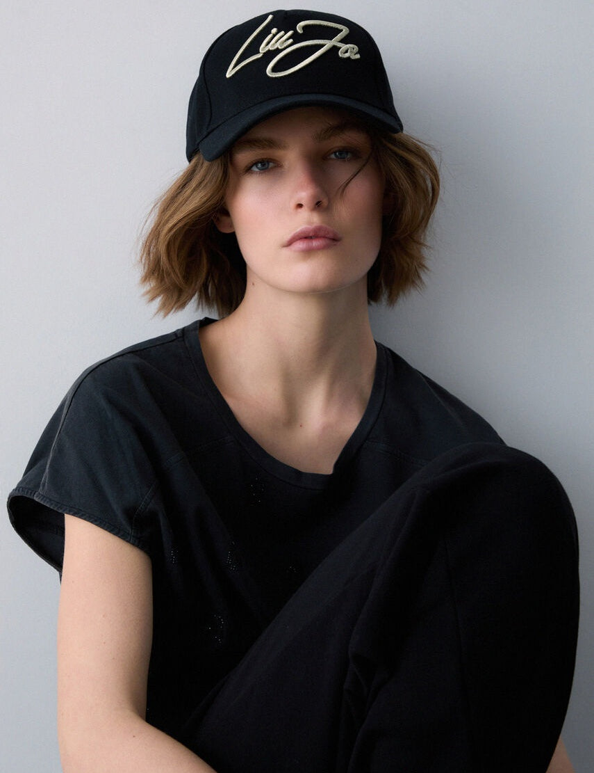 Modella con capelli corti e look total black indossa il Cappello baseball Liu Jo nero in canvas con logo oro ricamato sul davanti.