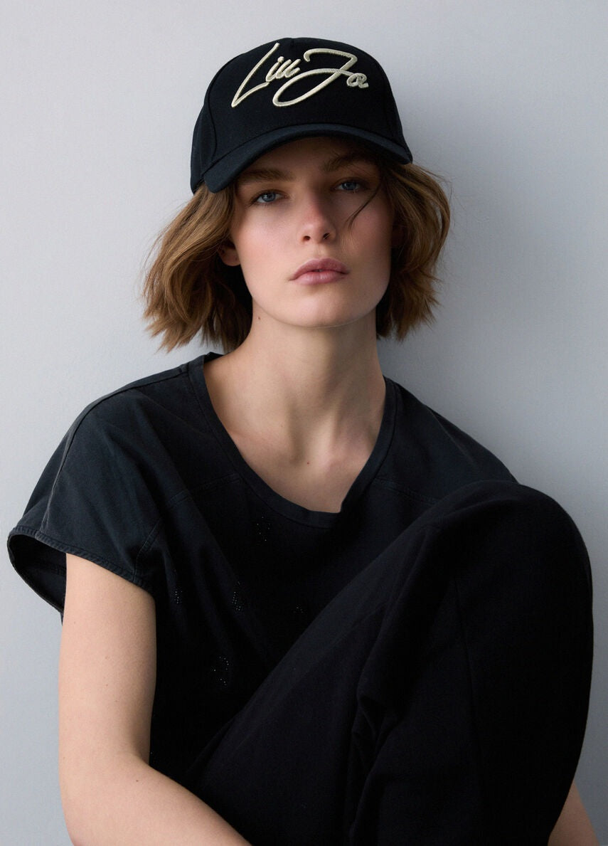 Modella con capelli corti e look total black indossa il Cappello baseball Liu Jo nero in canvas con logo oro ricamato sul davanti.